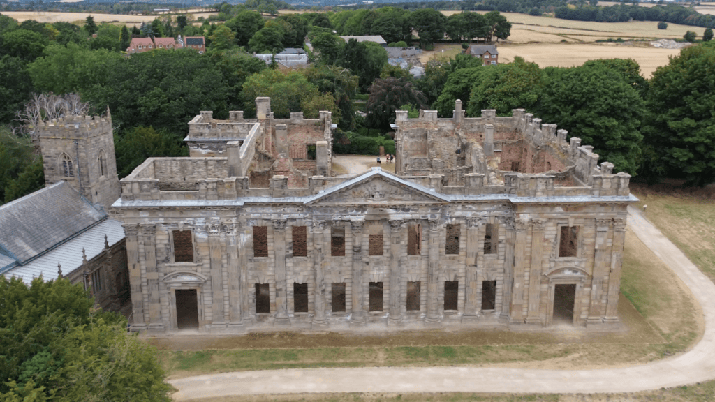 Aerial view of partially ruined historical building surrounded by trees and fields.