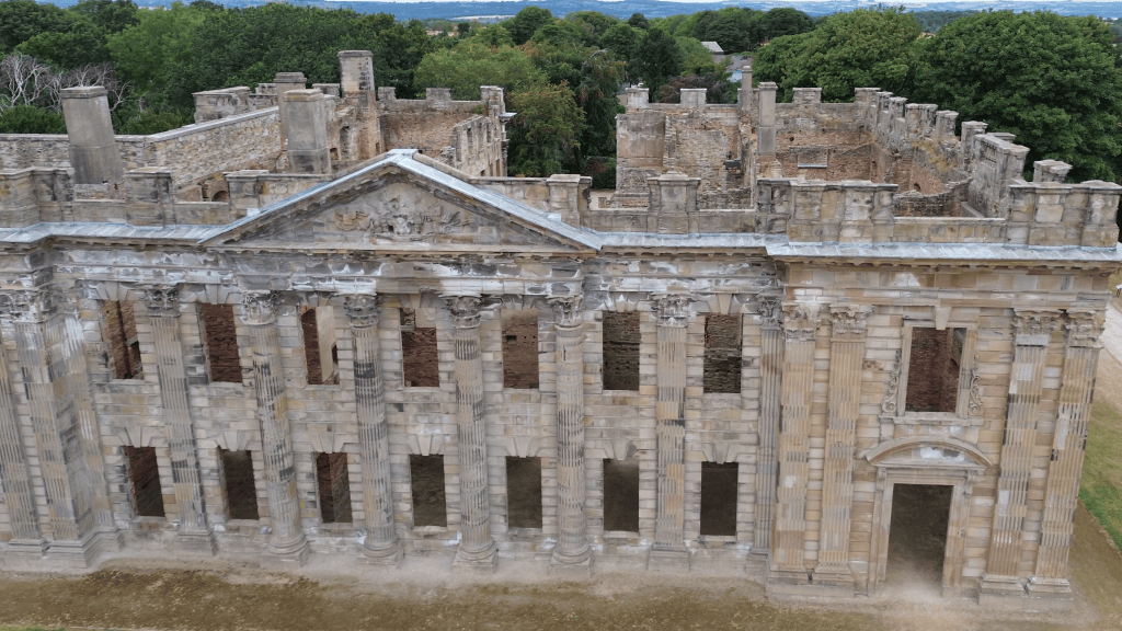 An aerial view of the ruins of an ornate stone building with classical architectural features, including tall columns and several broken windows, surrounded by greenery.