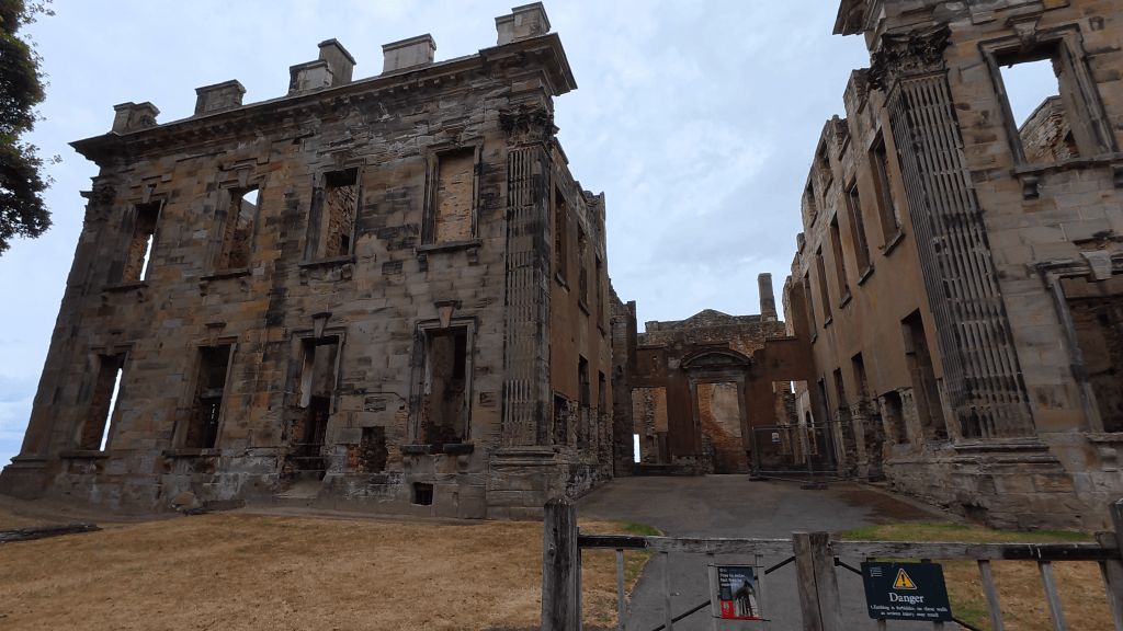 The ruins of a stone building with large windows and a grand architectural style, set against a cloudy sky.