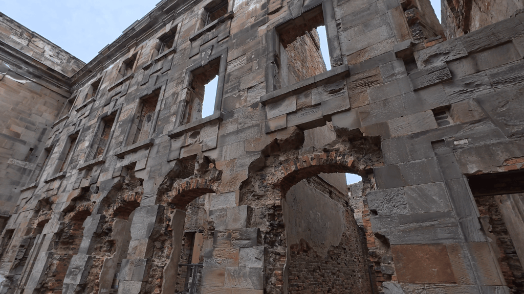 An abandoned building with crumbling stone walls, featuring several arched openings and large windows partially intact, under a cloudy sky.
